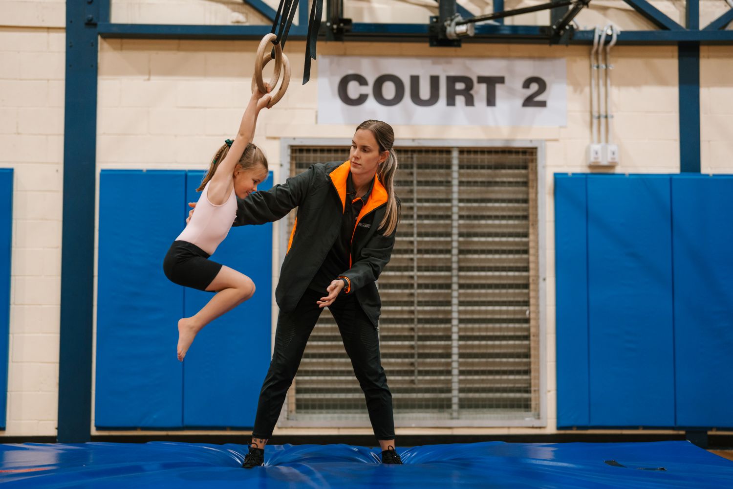 A female coach stands on a blue safety mat to spot a young girl hanging from wooden gymnastic rings in an indoor sports center marked "Court 2".